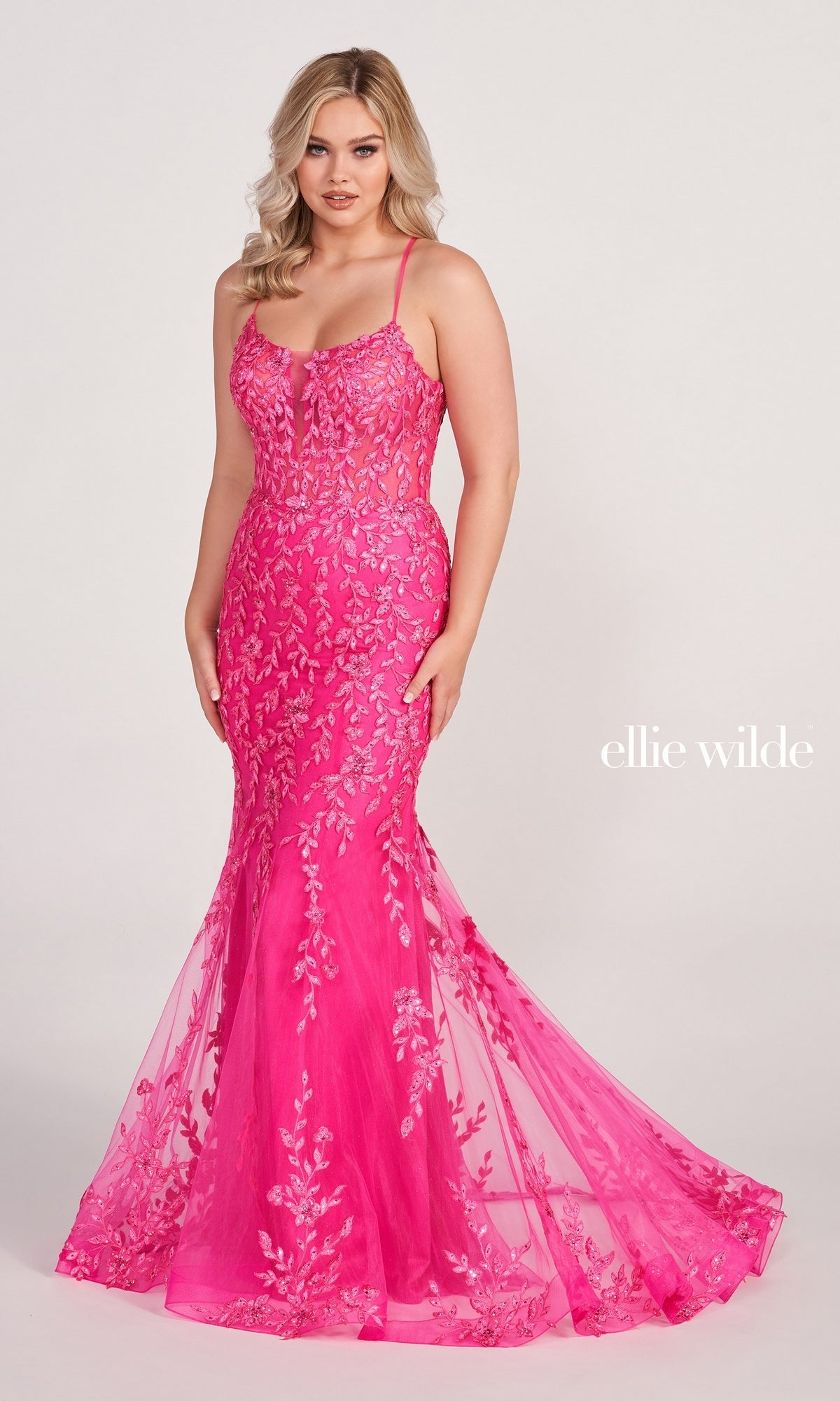 A woman wears the Ellie Wilde Lace Applique Over Glitter Jersey Gown EW34090—a fitted, floor-length bright pink dress with embroidered lace and thin straps—against a plain light background.